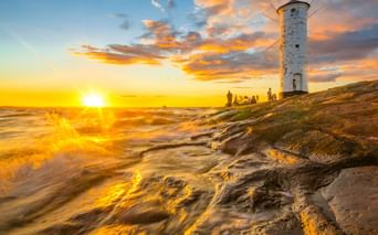 Weißer Windmühlen-Leuchtturm Mühlenbake in Swinemünde bei Sonnenuntergang. Goldene Wellen spülen über Sandstrand mit Silhouetten von Menschen.