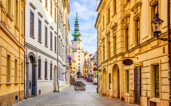 Schmale Kopfsteinpflasterstraße in Bratislavas Altstadt, gesäumt von gelben historischen Gebäuden. Ein Kirchturm mit grüner Kuppel ist im Hintergrund sichtbar.