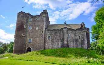 Doune Castle, eine mittelalterliche Steinfestung mit hohen Türmen und Mauern, thront auf einem grünen Hügel unter blauem Himmel mit weißen Wolken.