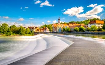 Historische Stadt Landsberg am Lech mit bunten Gebäuden und Kirchturm entlang des Lechs. Ein Wehr erzeugt fließendes Wasser im Vordergrund.
