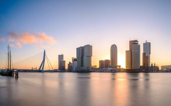 Rotterdamer Skyline bei Sonnenuntergang mit modernen Wolkenkratzern, die sich im ruhigen Wasser spiegeln. Die Erasmusbrücke links, Boote im Vordergrund.