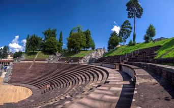 Antikes römisches Amphitheater von Augusta Raurica mit halbrunden Steinsitzreihen, grünem Hang und Bäumen unter blauem Himmel.