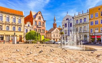 Kopfsteinpflasterplatz in der Altstadt von Tczew mit bunten historischen Gebäuden, Brunnen, Tauben und Kirchturm unter blauem Himmel.