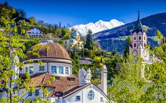Historisches Kurhaus mit Kuppeldach und Kirchturm in Meran, umgeben von grünen Bäumen mit schneebedeckten Alpengipfeln im Hintergrund.
