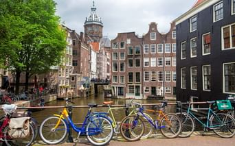 Reihe bunter Fahrräder entlang einer Amsterdamer Gracht mit traditionellen niederländischen Häusern und einem Kirchturm im Hintergrund unter bewölktem Himmel.