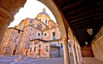 Blick durch einen Arkadenbogen auf den Dom von Mantua mit Backsteinfassade und blau gekachelter Kuppel. Das historische Gebäude wird von Steinsäulen gerahmt.