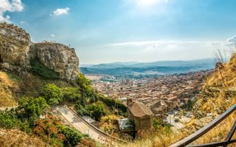 Panoramablick auf die Stadt Corleone in Sizilien mit historischen Festungsruinen auf Felsen, Terrakotta-Dächern und fernen Bergen unter blauem Himmel.