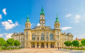 Gelbes barockes Rathaus in Győr mit drei grünen Kupfertürmen und verzierter Fassade. Menschen gehen über den Platz unter blauem Himmel.