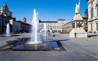 Piazza Castello in Turin mit hohen Springbrunnen im Vordergrund, weißem Denkmal rechts und historischen Palastgebäuden unter blauem Himmel.