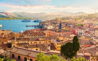 Luftaufnahme von Portoferraio mit bunten Gebäuden, Hafen mit Fähren und Bergen im Hintergrund unter weichem Abendhimmel.