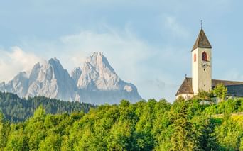 Weiße Kirche mit hohem Glockenturm in Innichen, umgeben von grünem Wald, mit dramatischen Dolomitengipfeln im Hintergrund unter blauem Himmel.