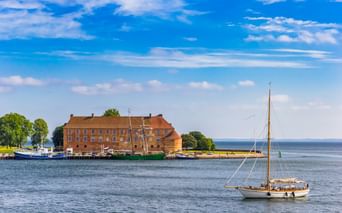 Schloss Sonderburg, ein Backsteingebäude der Renaissance, steht am Wasser mit einem Segelboot im Vordergrund und ruhigem blauen Wasser unter sonnigem Himmel.