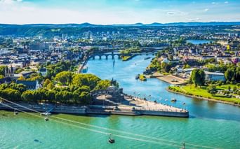 Luftaufnahme von Koblenz mit Deutsches Eck, wo zwei Flüsse zusammenfließen, Seilbahnen über dem Wasser und Festung Ehrenbreitstein im Vordergrund.