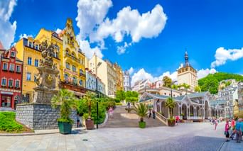 Karlsbad spa town square with colorful buildings, white colonnade, ornate fountain, and castle tower under blue sky with white clouds.