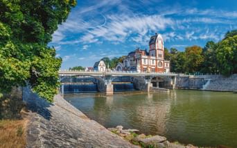 Steinbrücke über einen Fluss in Hradec Králové mit einem Uhrturm aus rotem Backstein. Grüne Bäume säumen die Flussufer unter blauem Himmel.