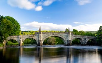 General Wade's Bridge, eine historische Steinbrücke mit fünf Bögen über einem Fluss in Aberfeldy. Üppige grüne Bäume rahmen beide Seiten unter blauem Himmel.