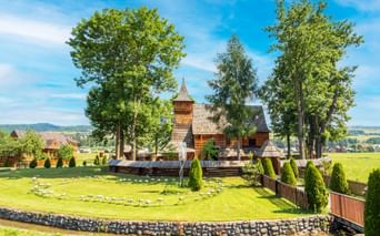 Traditionelle Holzkirche in Dąbno mit Schindeldach und Glockenturm, umgeben von hohen Bäumen und gepflegtem Rasen mit kreisförmiger Steinumrandung.