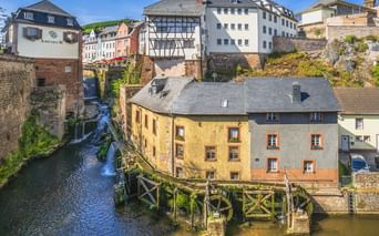 Wasserfall fließt durch Saarburgs Altstadt mit traditionellen Fachwerkhäusern, Steinmauern und einer Holzbrücke über dem Bach.
