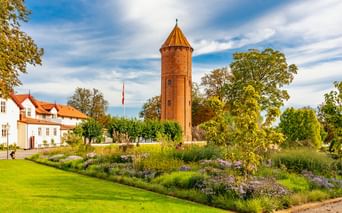 Roter Backsteinwasserturm in Praestø mit Kegeldach, umgeben von Gartenbeeten und grünem Rasen. Weißes Gebäude mit rotem Dach links sichtbar.