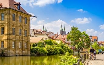 Radfahrer am Flussufer in der Bamberger Altstadt mit historischem Sandsteingebäude, bunten Häusern und Doppelkirchtürmen unter blauem Himmel.