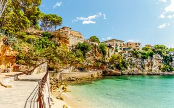 Malerische Bucht in Porto Cristo mit türkisfarbenem Wasser, Sandstrand und Küstenpromenade. Steingebäude und Hotels auf Felsenklippen mit Pinien.