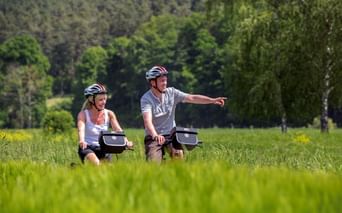 Zwei Radfahrer mit Helmen fahren durch grüne Wiese im Altmühltal. Mann zeigt nach vorne, Wald im Hintergrund. Sonniger Sommertag.