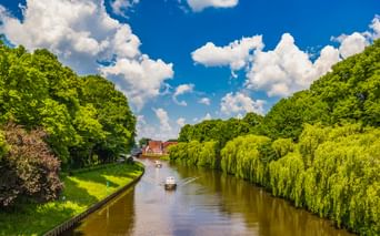 Fluss Ems in Meppen fließt zwischen üppig grünen Bäumen und Weiden. Zwei Boote fahren auf dem ruhigen Wasser unter blauem Himmel mit weißen Wolken.