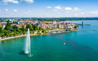 Luftaufnahme von Friedrichshafen am Bodensee mit hoher Fontäne im türkisfarbenen Wasser, historischer Altstadt, Kirchturm und Fahrgastschiff.