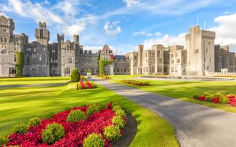 Ashford Castle in Irland mit Steintürmen und Türmchen. Gepflegte Rasenflächen mit roten und grünen Blumenbeeten säumen die Auffahrt unter blauem Himmel.