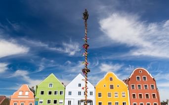 Hoher geschmückter Maibaum vor bunten Giebelhäusern in Obernberg am Inn. Gebäude in Grün, Weiß, Gelb und Rot unter blauem Himmel.