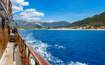 Blick vom Holzbootsdeck auf blaues Wasser nahe Lefkada. Berge und Küstenstadt in der Ferne unter klarem Himmel sichtbar.
