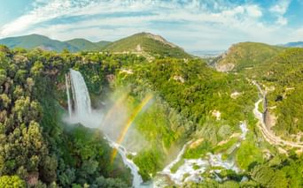 Luftaufnahme des Wasserfalls Cascata delle Marmore in Italien mit Regenbogen im Nebel, umgeben von grünen Bergen und Serpentinenstraße.
