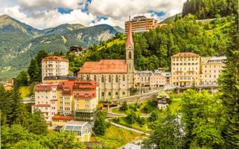 Pfarrkirche mit hohem Turm und rotem Dach in Bad Gastein, umgeben von bunten Gebäuden am Hang mit bewaldeten Bergen im Hintergrund.