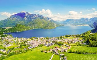 Luftaufnahme von St. Gilgen mit grünen Wiesen am Wolfgangsee, umgeben von bewaldeten Bergen unter blauem Himmel in Österreich.