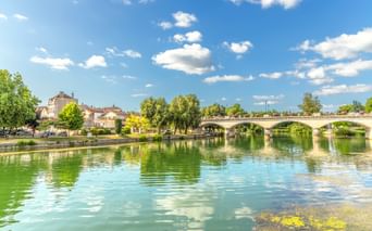 Charente-Fluss in Cognac mit ruhigem grünen Wasser, das Wolken spiegelt. Steinbrücke mit mehreren Bögen überspannt den Fluss, gesäumt von Bäumen.