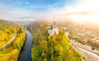 Luftaufnahme der Burg Rosenberg auf einem bewaldeten Hügel, umgeben von einer Flussschleife. Herbstbäume in goldenen Farben, Morgennebel und Sonnenlicht.