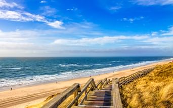 Holztreppe führt durch Dünengras zum Sandstrand in Wenningstedt, Sylt. Blauer Himmel mit weißen Wolken über der Nordsee.