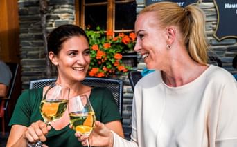 Zwei lächelnde Frauen stoßen mit Weißweingläsern auf einer Außenterrasse an. Orangefarbene Blumen und Weinfässer im Hintergrund.