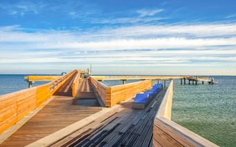 Moderne Holzsteganlage mit blauen Bänken, die in die Ostsee in Heiligenhafen hineinragt, unter blauem Himmel mit vereinzelten Wolken.