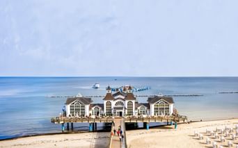 Historische Seebrücke Sellin in der Ostsee mit weißem Pavillongebäude, Sandstrand mit Strandkörben und ruhigem blauem Wasser.