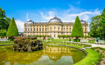 Würzburger Residenz mit gelber Barockfassade und Kuppel, vom formalen Garten mit Zierteich und kegelförmigen Bäumen aus gesehen.