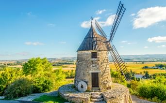 Steinwindmühle mit blauem Kegeldach und Holzflügeln in Castelnaudary, mit Blick auf hügelige Landschaft mit Feldern unter blauem Himmel.