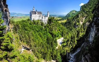Schloss Neuschwanstein thront auf einem Hügel, umgeben von dichtem Wald und Bergen. Eine Schlucht mit Felsklippen rahmt die Ansicht in Bayern ein.
