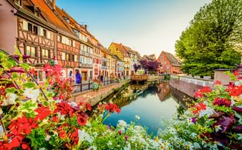 Kanal gesäumt von bunten Fachwerkhäusern in Colmar, Frankreich. Rote und weiße Blumen rahmen die Ansicht, mit Bäumen und blauem Himmel.