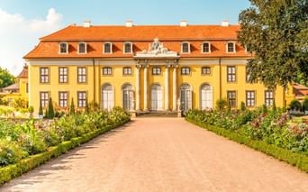 Yellow baroque palace with orange roof and white columns in Mosigkau near Dessau. A gravel path leads through a formal garden to the entrance.