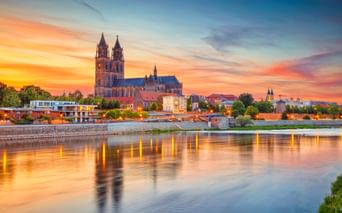 Magdeburger Dom mit Doppeltürmen an der Elbe bei Sonnenuntergang. Farbenfroher Himmel spiegelt sich im ruhigen Wasser, Stadtgebäude sichtbar.