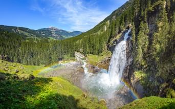 Krimmler Wasserfälle stürzen über Felsklippen mit Regenbogen im Nebel, umgeben von grünen Almwiesen und bewaldeten Bergen in Österreich.