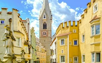 Bunte mittelalterliche Gebäude mit Zinnen säumen eine enge Gasse in Brixens Altstadt, mit einem weißen Kirchturm in der Mitte.