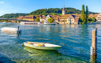Blick auf Stein am Rhein am Rheinufer mit Booten im Vordergrund, historischen Gebäuden, Kirchturm und bewaldeten Hügeln im Hintergrund.