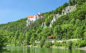 Burg Prunn thront auf weißen Kalksteinfelsen über grünen Wäldern bei Riedenburg. Die Altmühl fließt im Vordergrund.